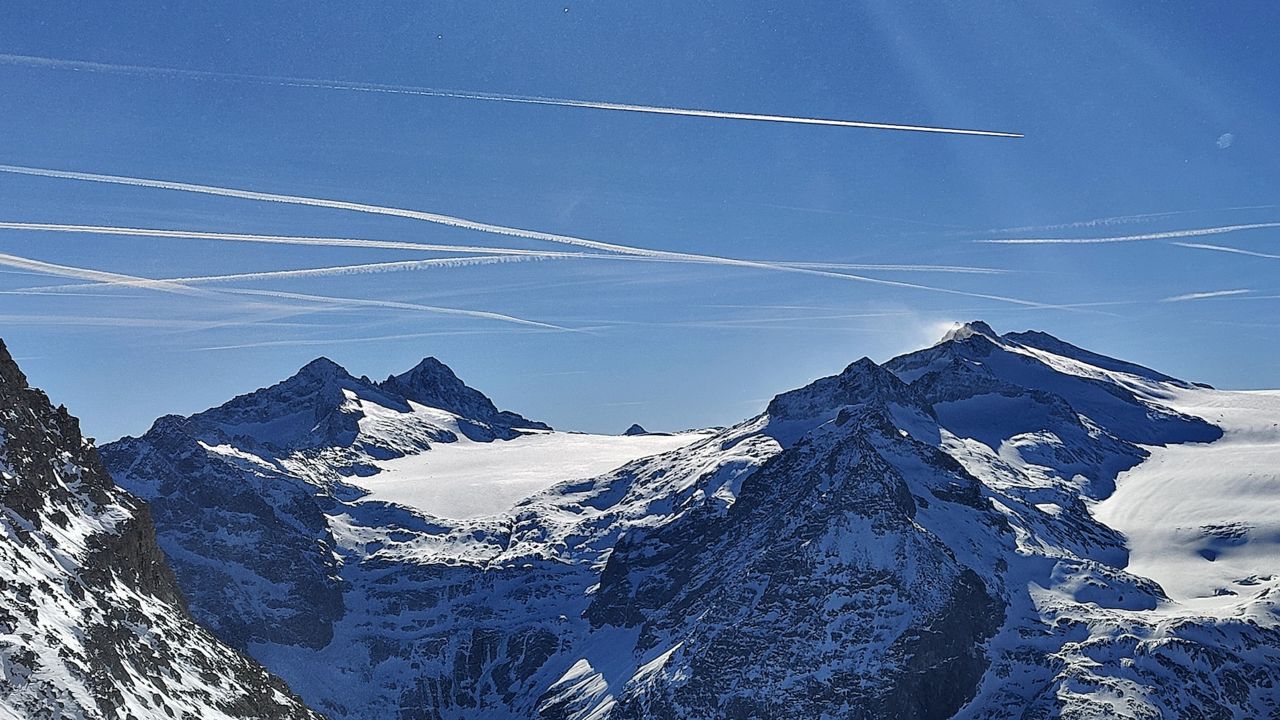 Die Presena-Gletscher in den Alpen. Man sieht die Gipfel der Bergkette, die von Eis überzogen sind und die Spuren der Flugzeuge im Himmel dahinter.