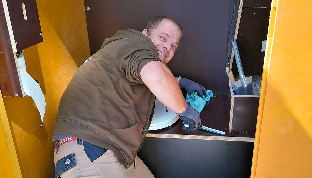Unser Mitarbeiter "Hüne", der gerade eine unserer Kabinen reinigt. Er ist über die Sitzfläche gebeugt, zieht die Fläche gerade mit einem Reinigungsschrubber ab und lächelt dabei über seine Schulter in die Kamera.
